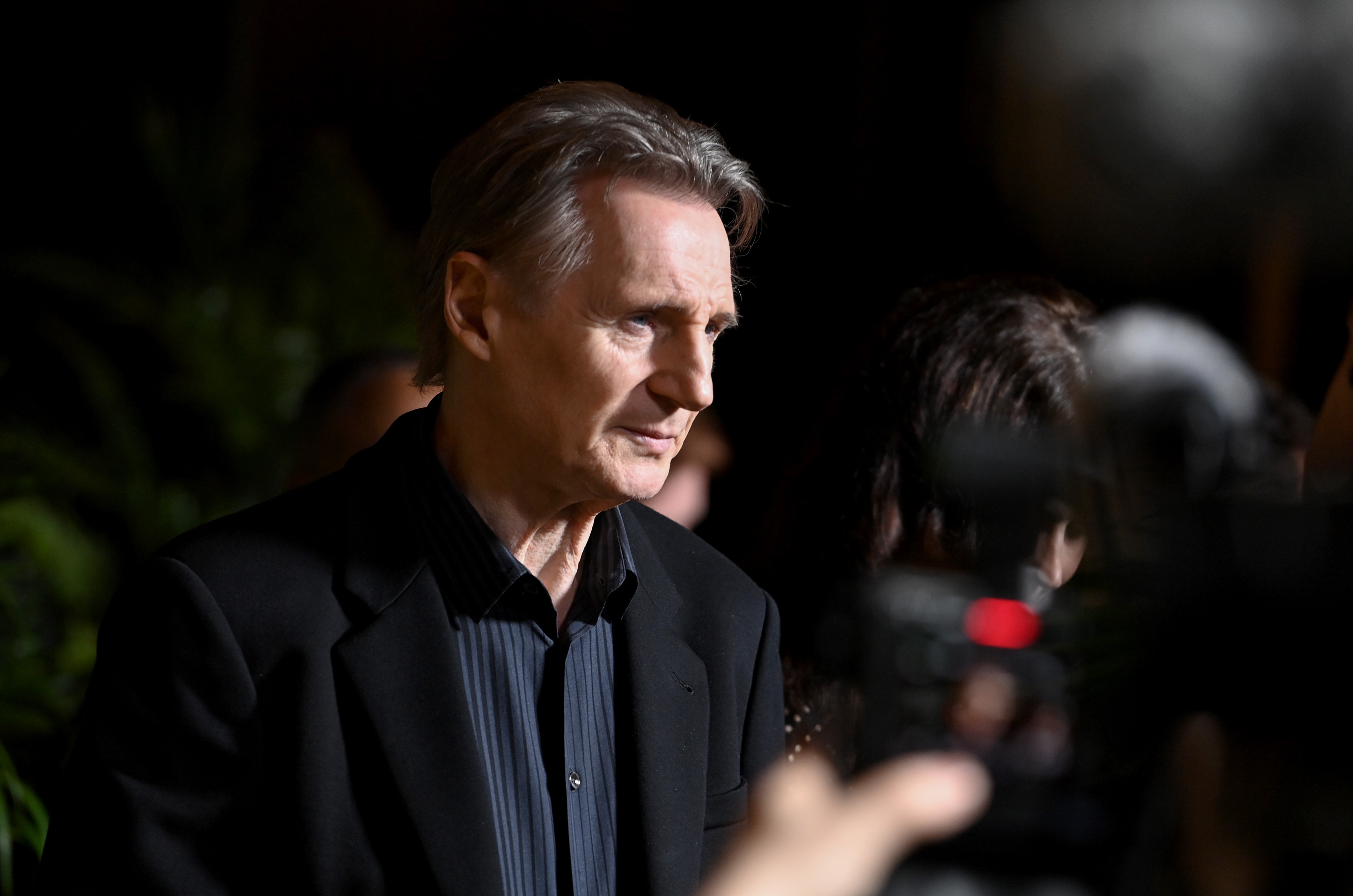 Liam Neeson successful  a acheronian  suit   stands astatine  a nationalist   event, facing cameras and radical   nearby