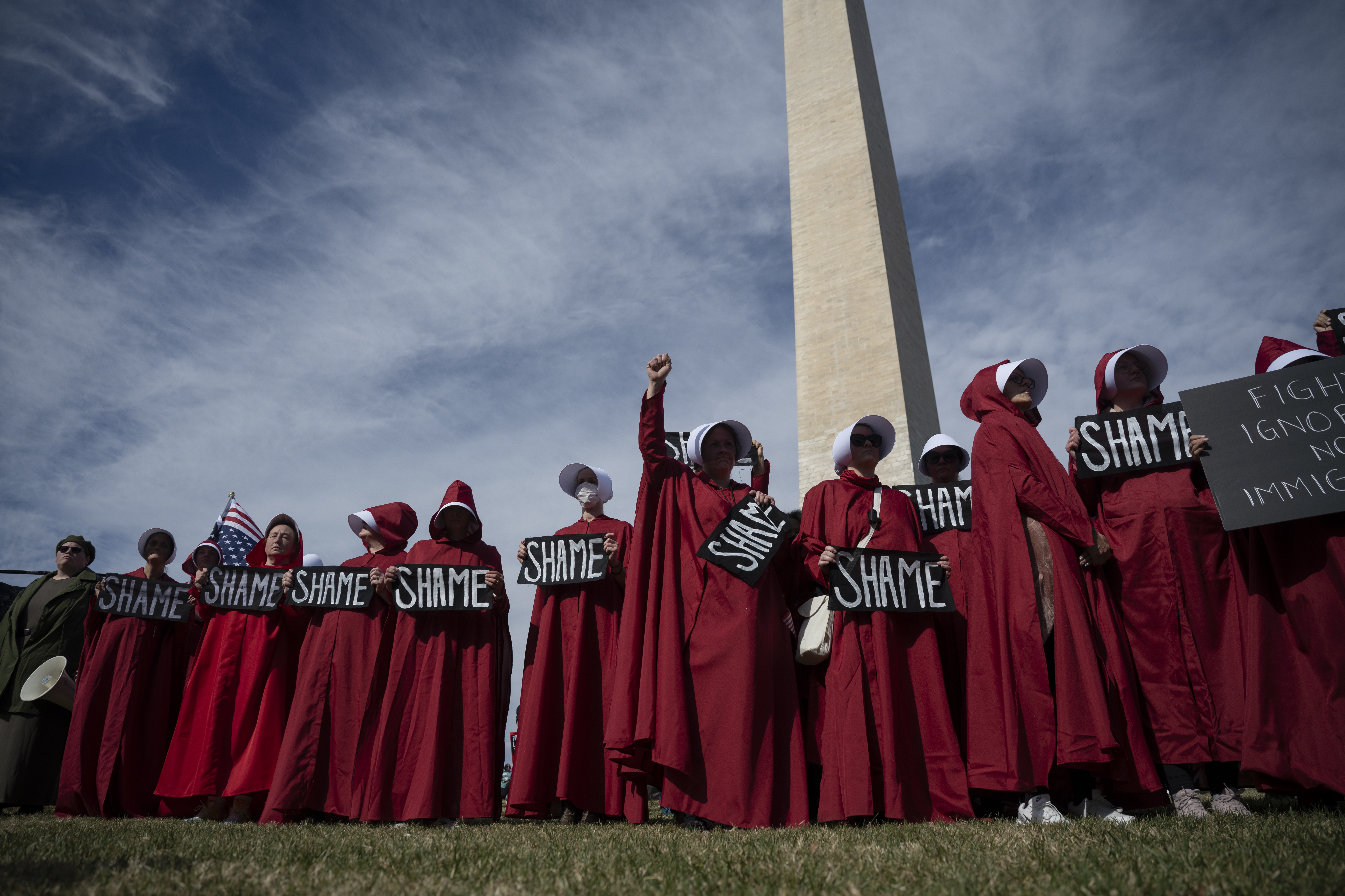En gruppe kledd i røde kapper og hvite panser holder «Skam»; skilt, protesterer nær et høyt monument under en overskyet himmel