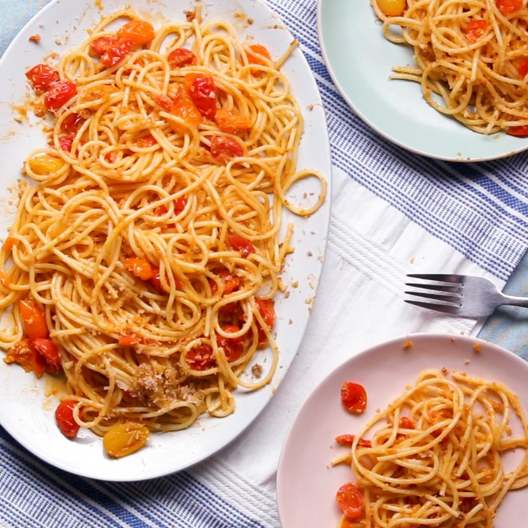 Platter and plates of spaghetti with cherry tomatoes and breadcrumbs, placed on a striped tablecloth