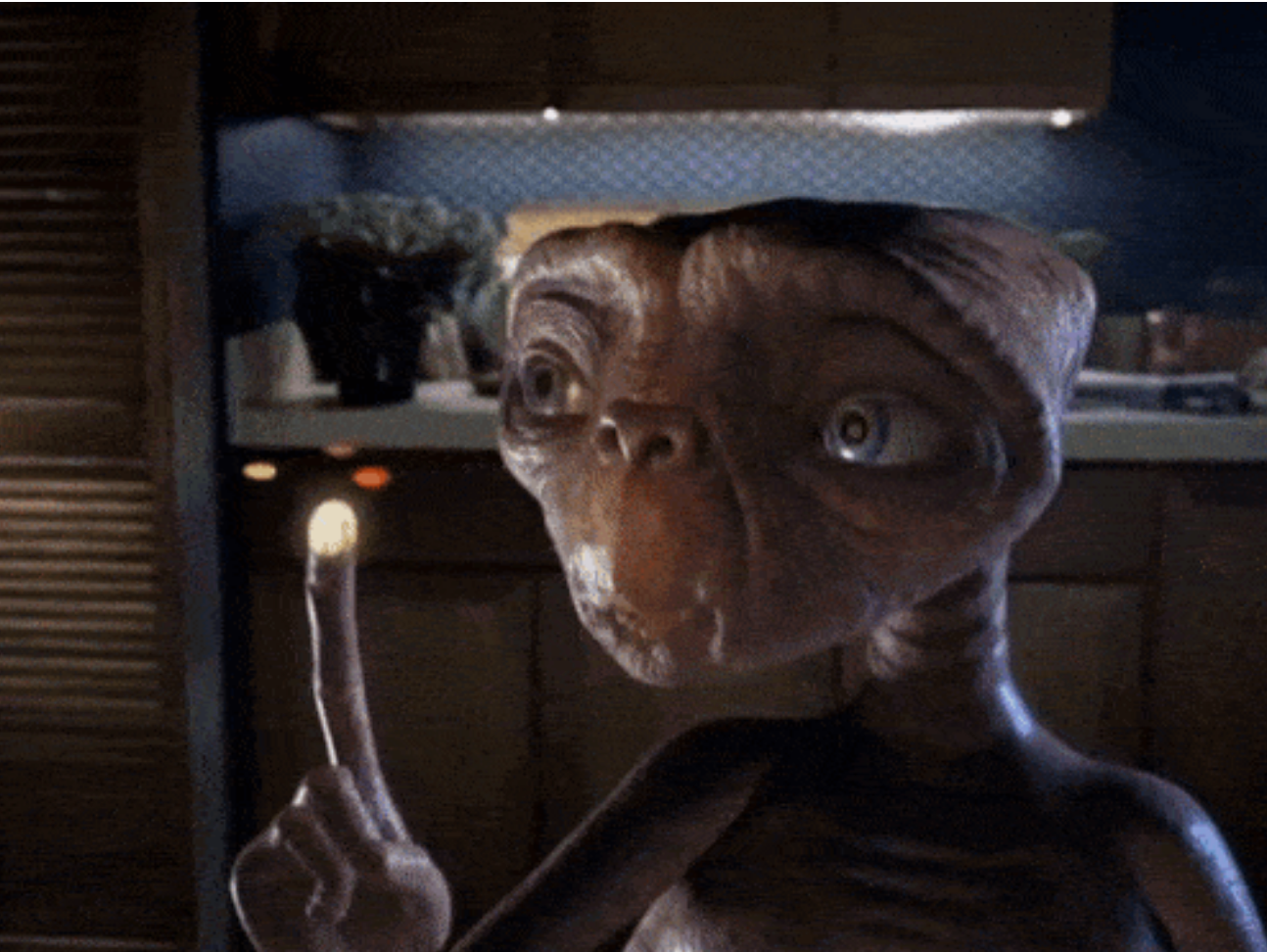 An alien with a glowing fingertip points upward, in a kitchen setting