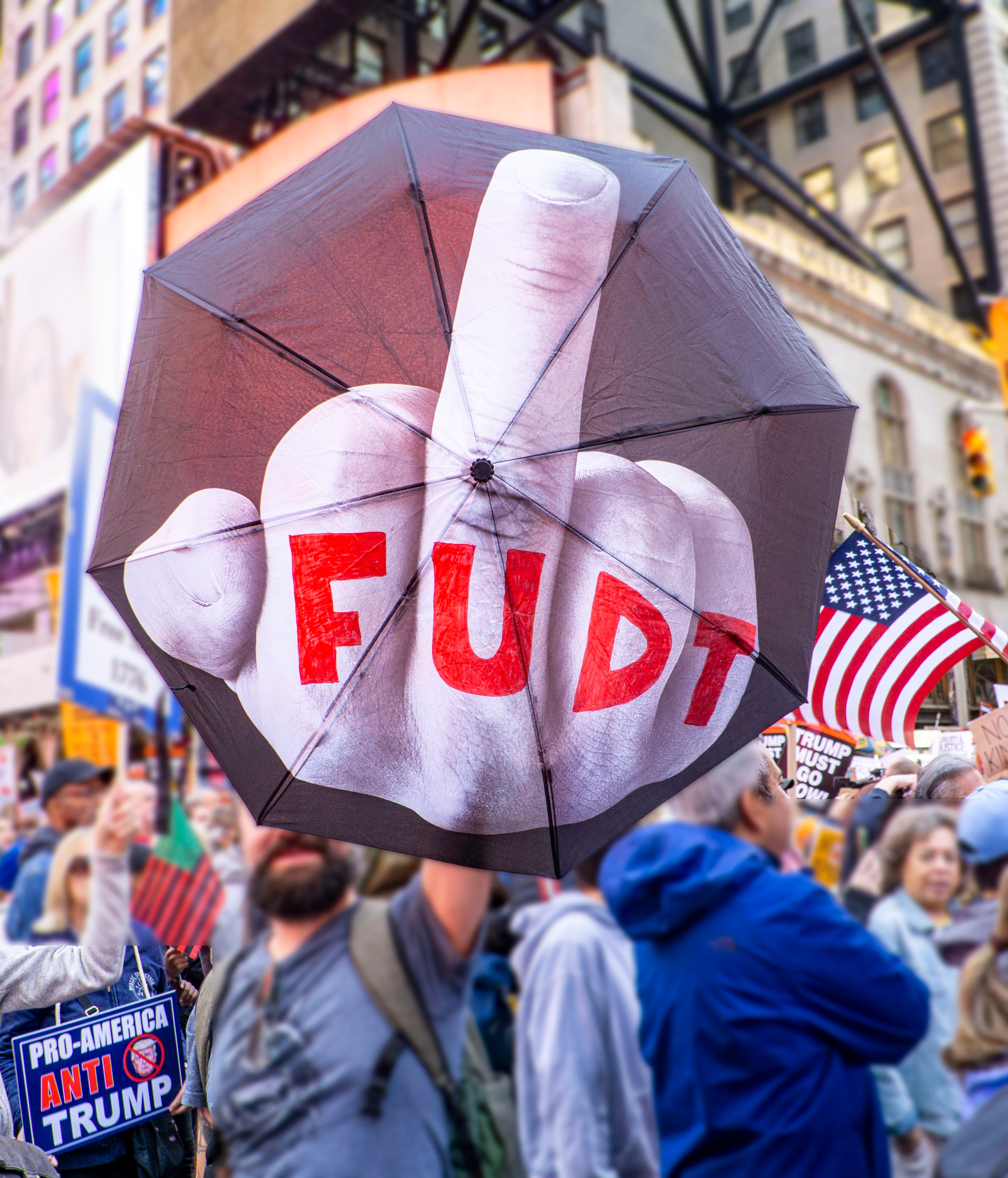 Person holder en paraply med en uttrykksfull håndbevegelse og "FU DT" tekst ved en anti-Trump-protest. Amerikansk flagg og protestskilt synlig