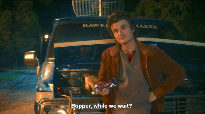 Person leaning on an open van hood, holding a chocolate bar. Subtitle reads: "Bopper, while we wait?"