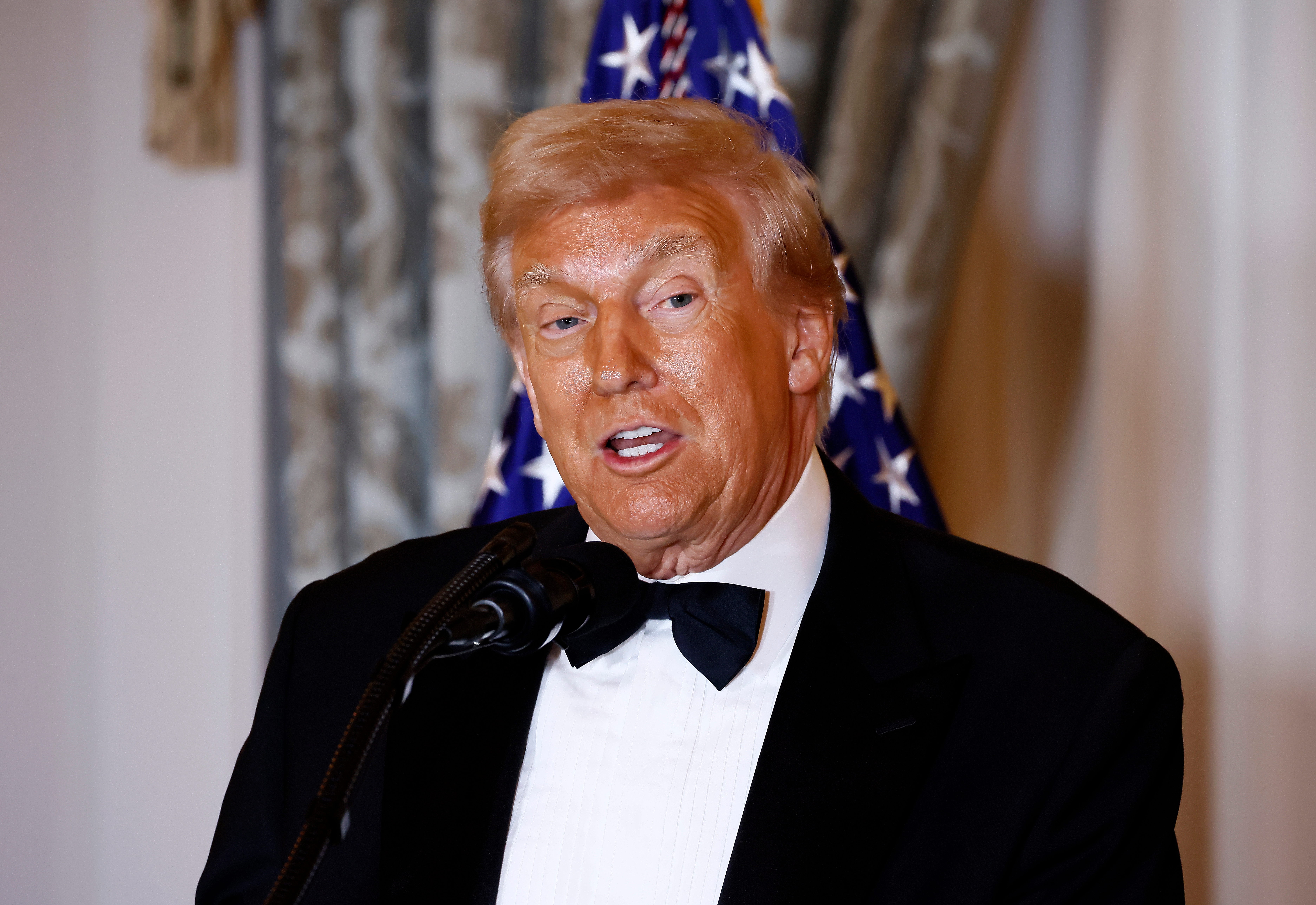 Mann i smoking snakker på et podium med et amerikansk flagg i bakgrunnen, og gestikulerer lett