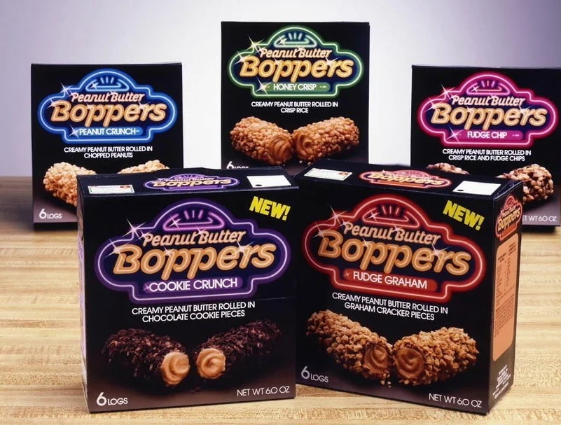 Five boxes of Peanut Butter Boppers in various flavors are displayed on a wooden surface. Each box shows an image of the snack inside