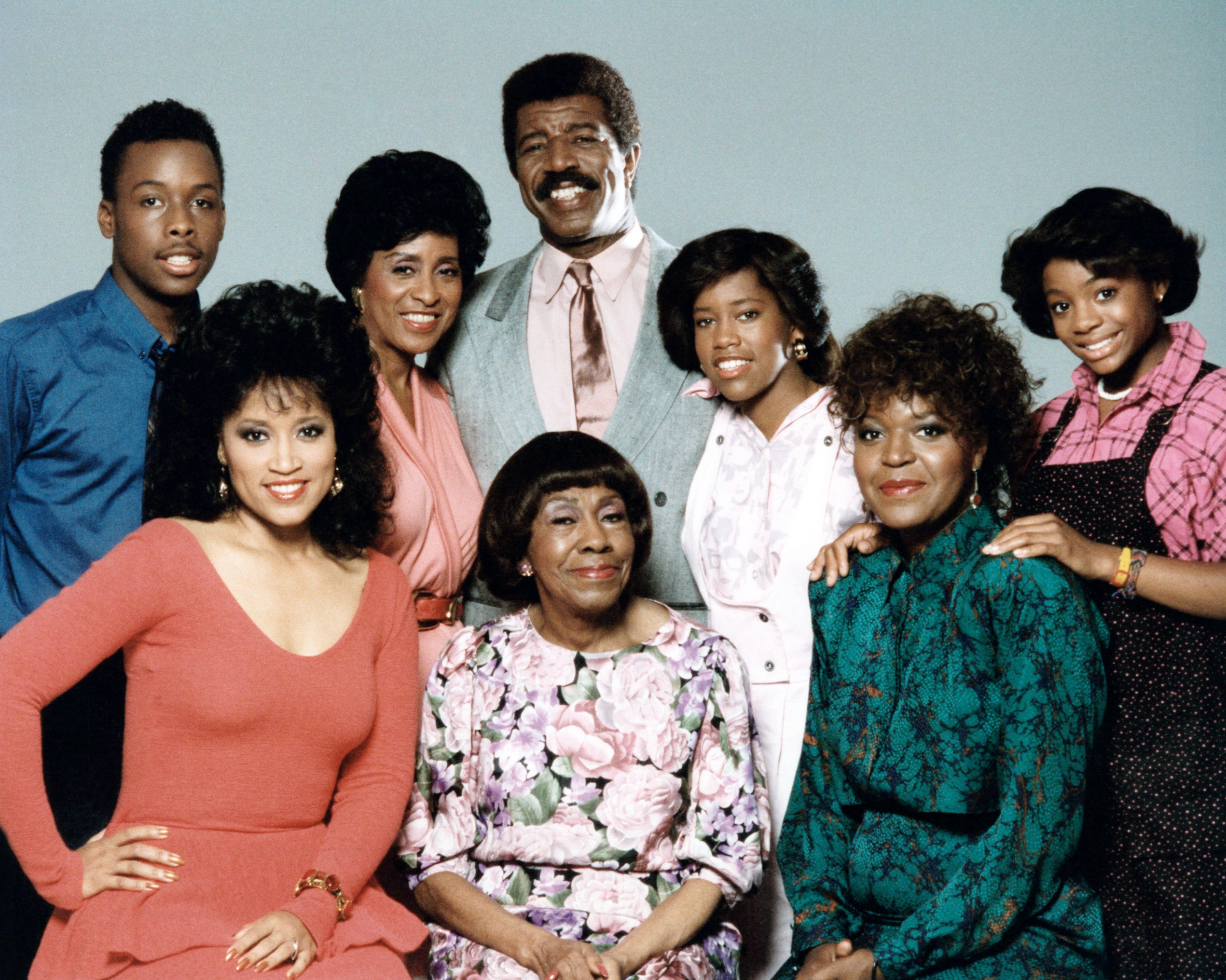 A retro family portrait with eight people dressed in 1980s fashion, including suits and patterned dresses, smiling at the camera