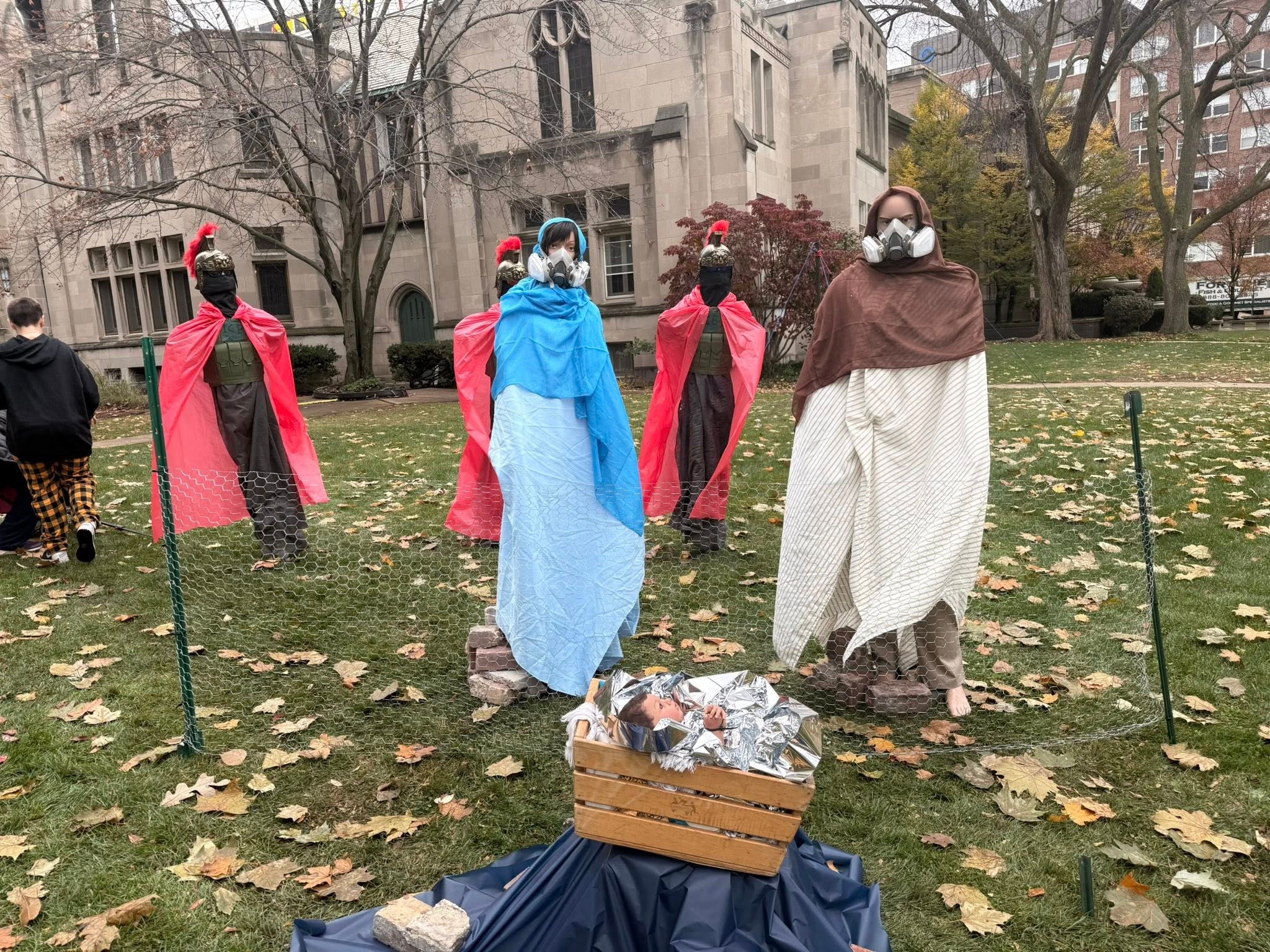 Utendørs julekrybbe med tre skikkelser iført ansiktsmasker og kapper, omgitt av løv. En kurv foran representerer en krybbe