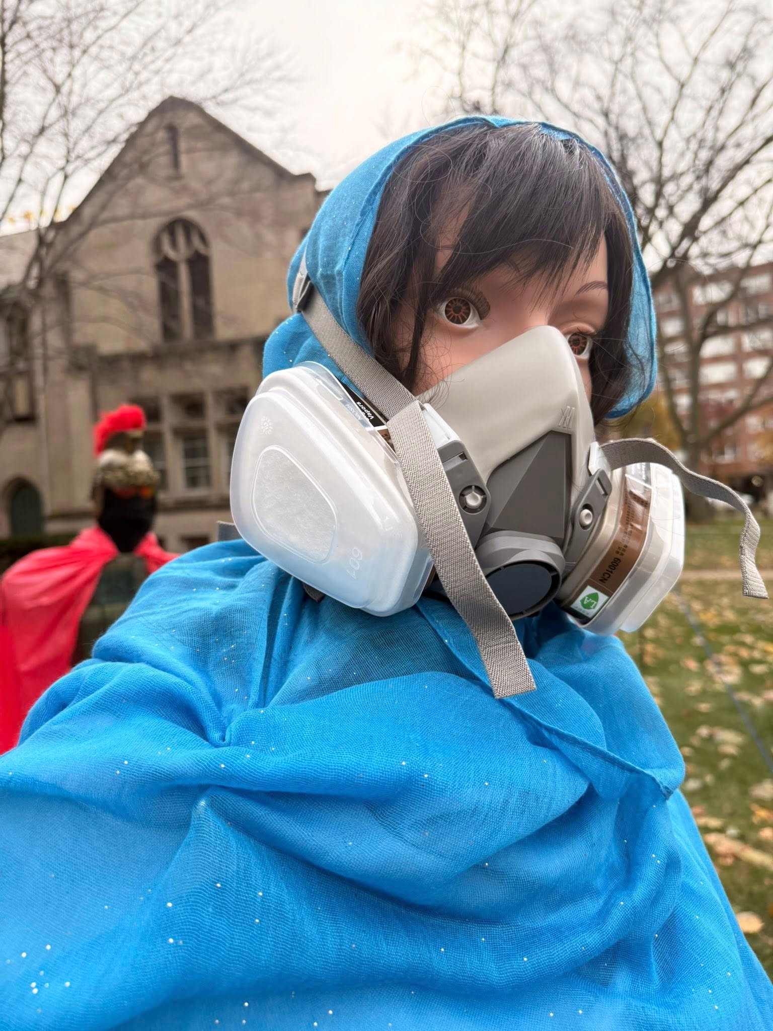 En utstillingsdukke i et blått skjerf og åndedrettsmaske står utendørs, med en uskarp bakgrunn inkludert en bygning og en annen mannequin