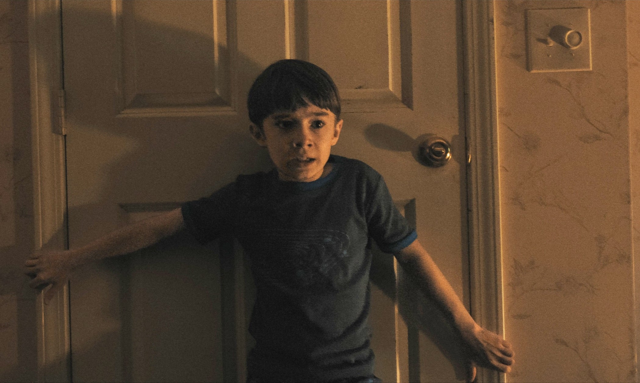 یک کودک در اتاق تاریک مقابل در بسته ایستاده، ترسیده و دستگیره در را می‌گیرد
