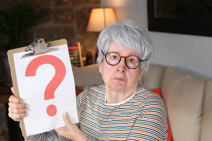 Person with glasses and striped shirt holding a clipboard with a large red question mark, sitting on a couch in a room with a lamp