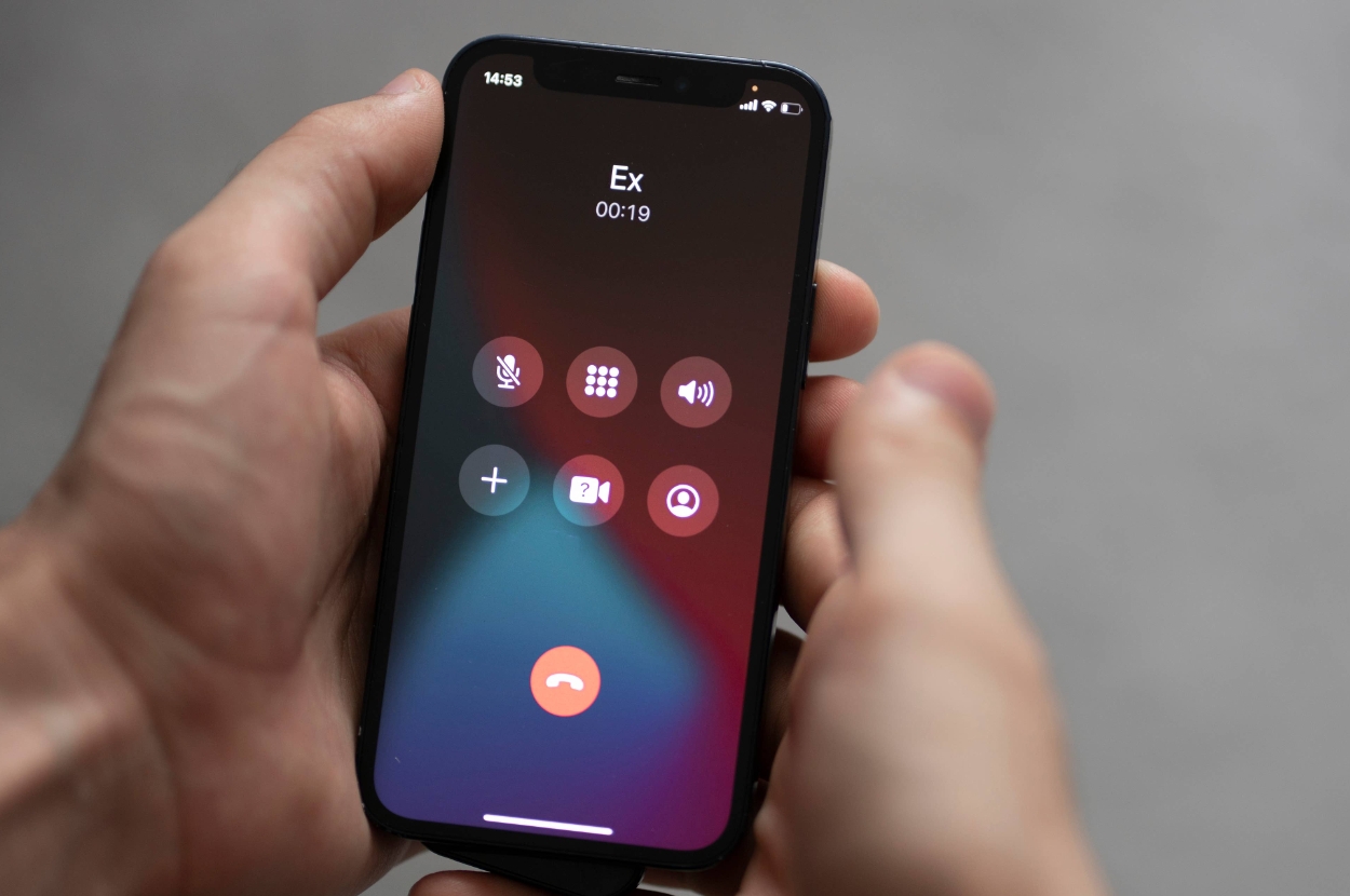 Person holding a smartphone displaying an active call with "Ex," showing call duration and icons for mute, keypad, speaker, FaceTime, and add call