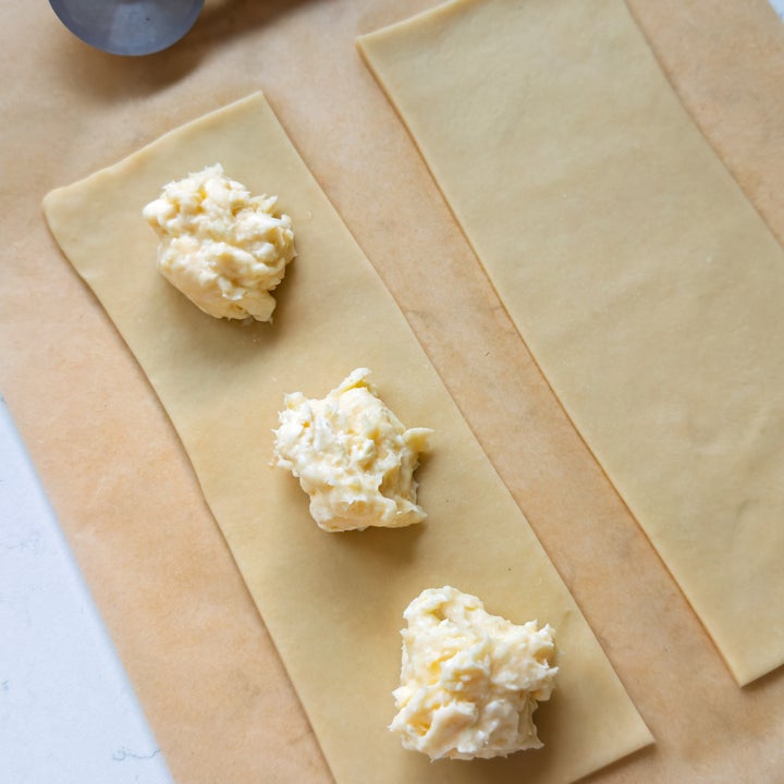 Two sheets of pasta dough on parchment paper with dollops of ricotta cheese filling, next to a pasta cutter