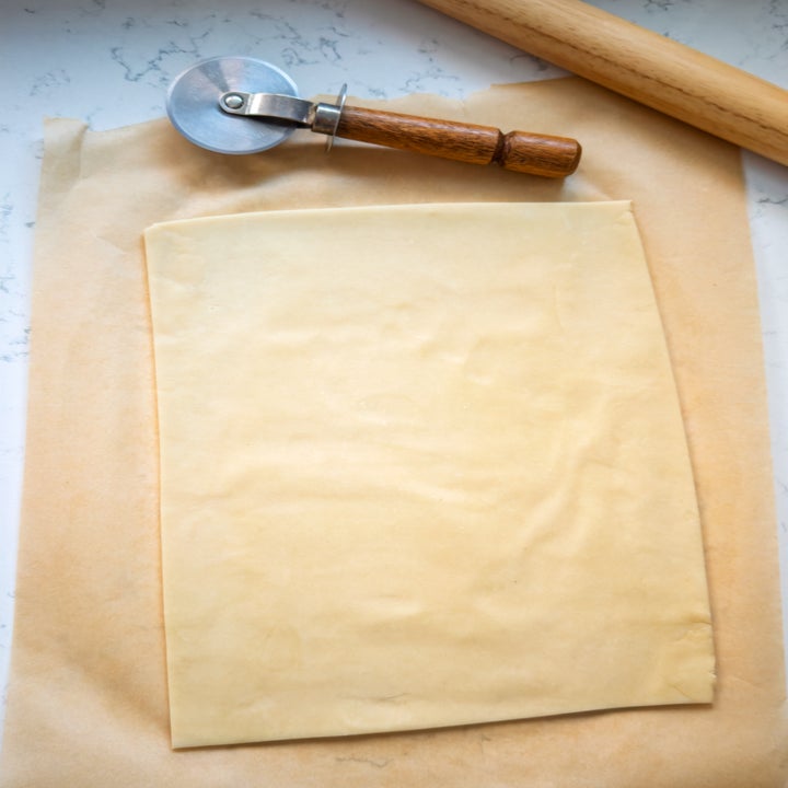 Unbaked rectangular pastry dough on parchment paper, next to a wooden-handled pizza cutter and rolling pin