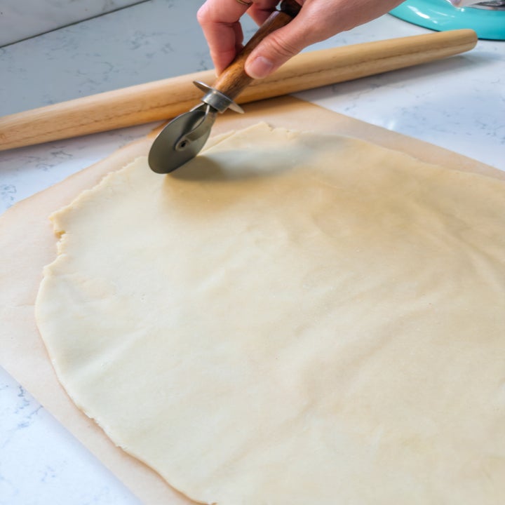 Hand holding a pizza cutter, about to cut rolled dough on parchment, with a rolling pin nearby on a countertop