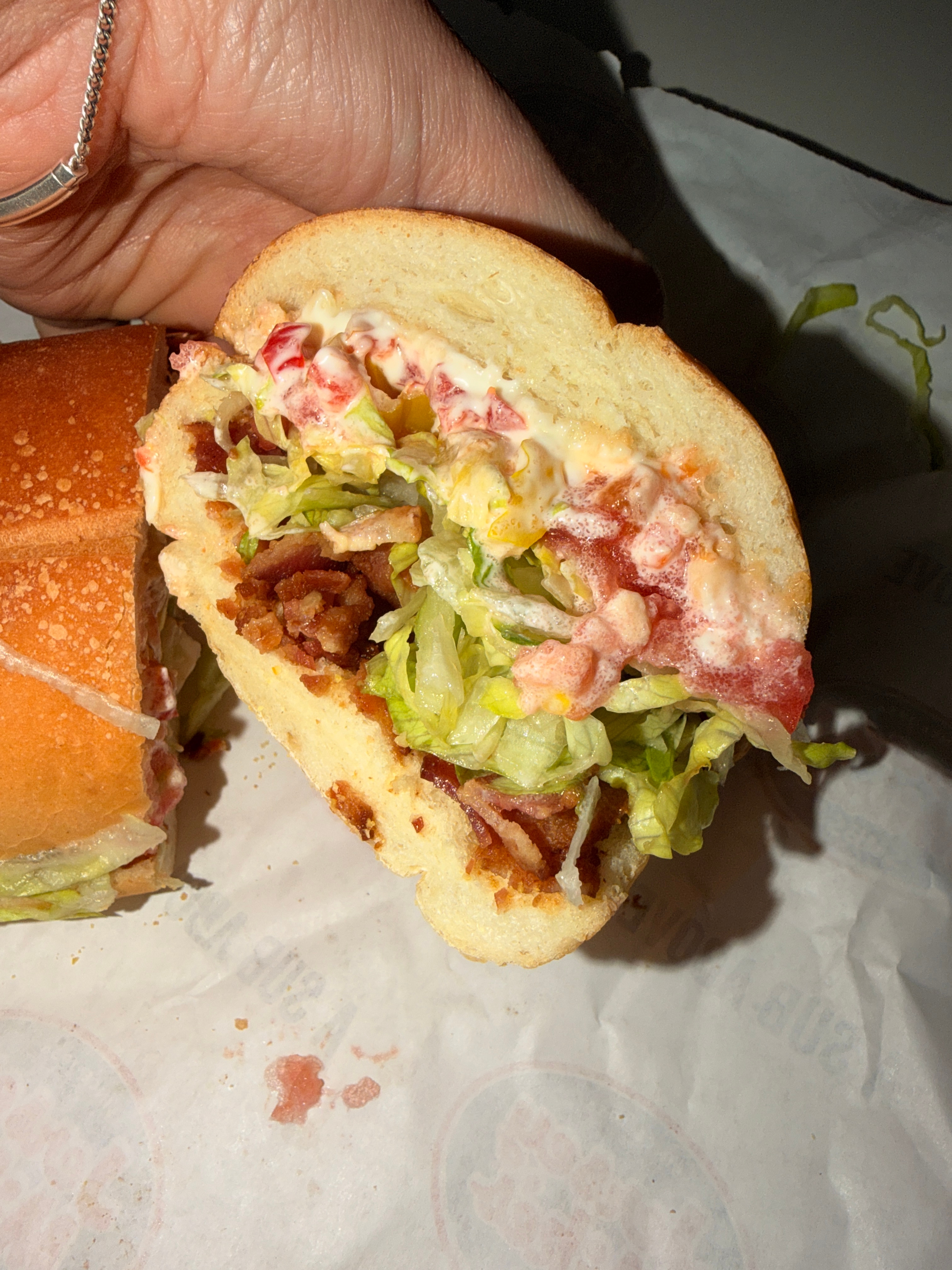A hand holds a sandwich filled with crispy bacon, lettuce, tomato, and mayonnaise on a bread roll