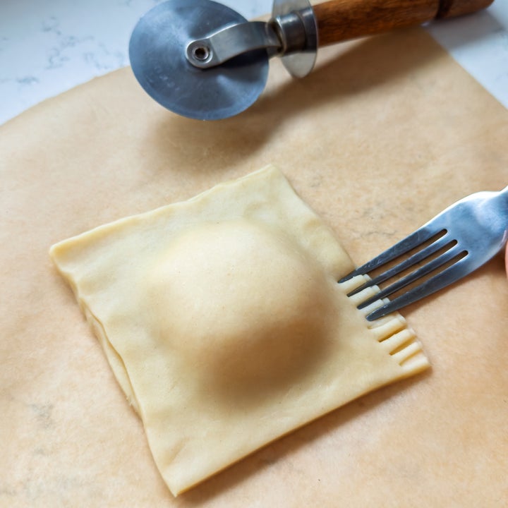 A square ravioli piece is being sealed with a fork on parchment paper, next to a pizza cutter