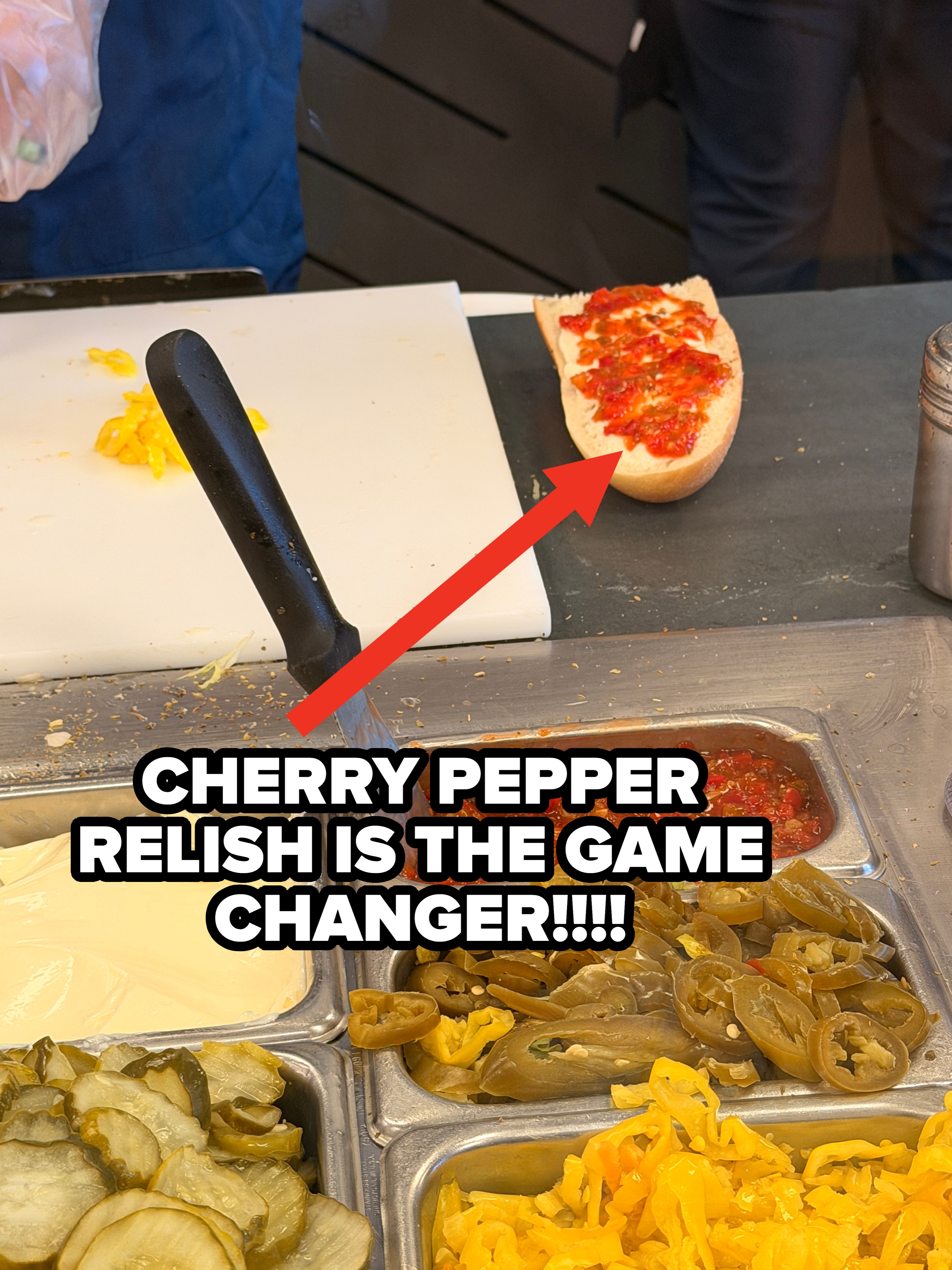 Sandwich station with sliced pickles, jalapeños, banana peppers, mayonnaise, chopped chilies, and an open sandwich being prepared