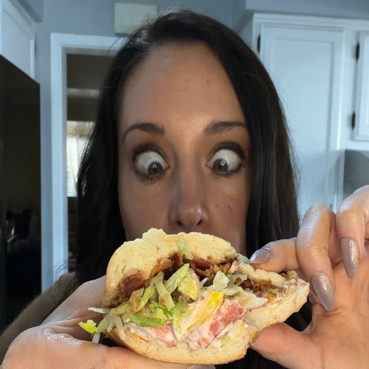A woman with a surprised expression holds a sandwich filled with lettuce, tomatoes, and bacon, ready to take a bite in a kitchen setting