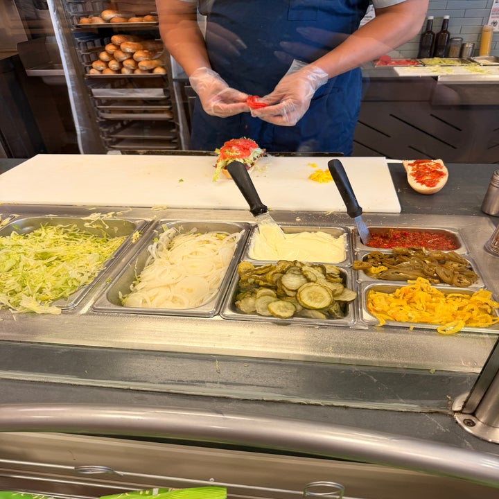 Person in gloves prepares a sandwich at a deli counter with toppings like lettuce, onions, pickles, and jalapeños visible in trays