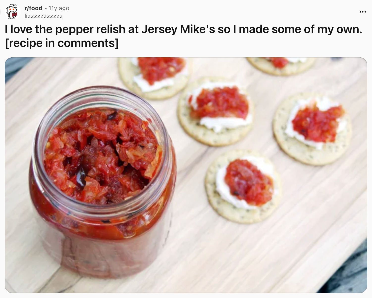 Jar of homemade pepper relish with crackers topped with cream and relish spread on a wooden board. Recipe mentioned in comments