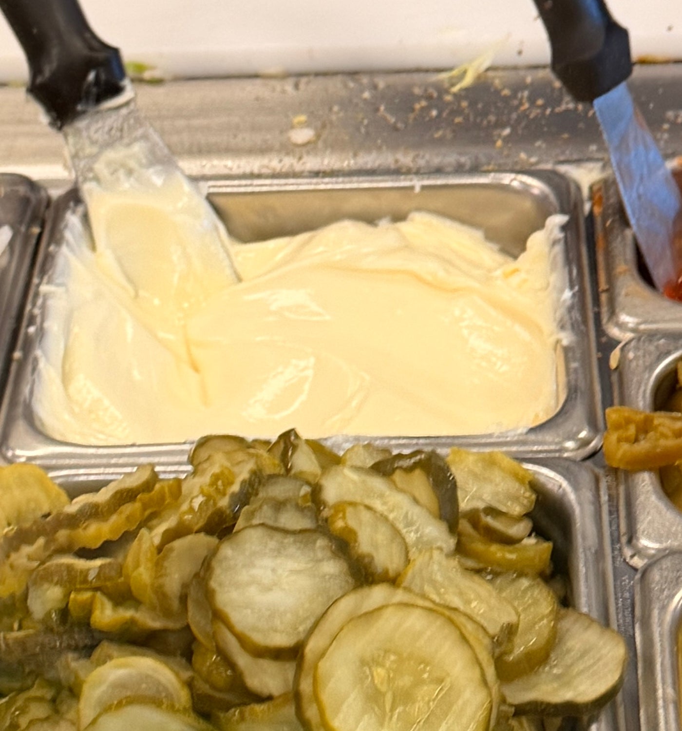 Sandwich shop prep station with trays of onions, pickles, shredded cheese, and sauces. Chef prepares a sandwich in the background