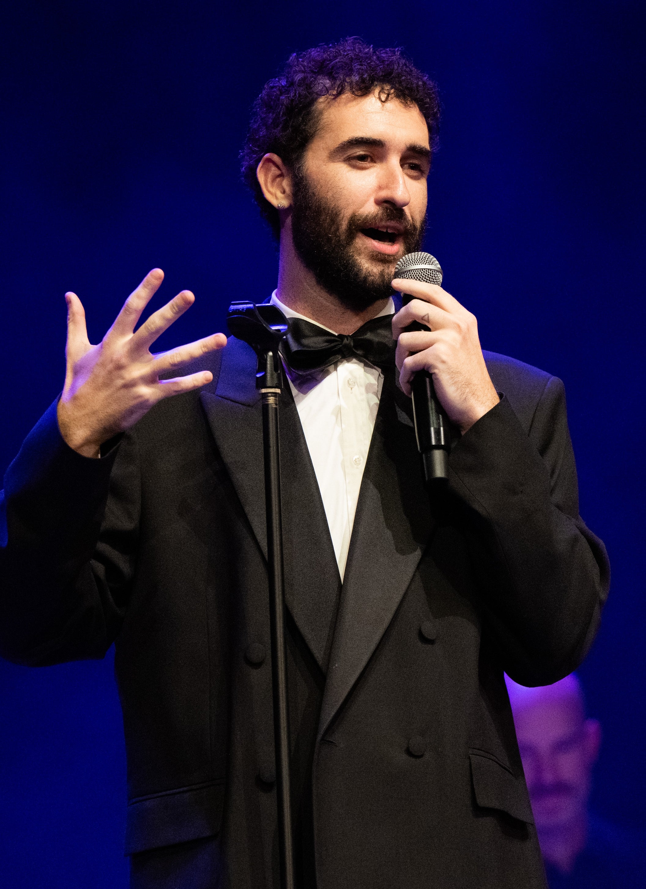 A idiosyncratic successful a suit overgarment and bow necktie speaks into a microphone connected stage, gesturing expressively with 1 hand