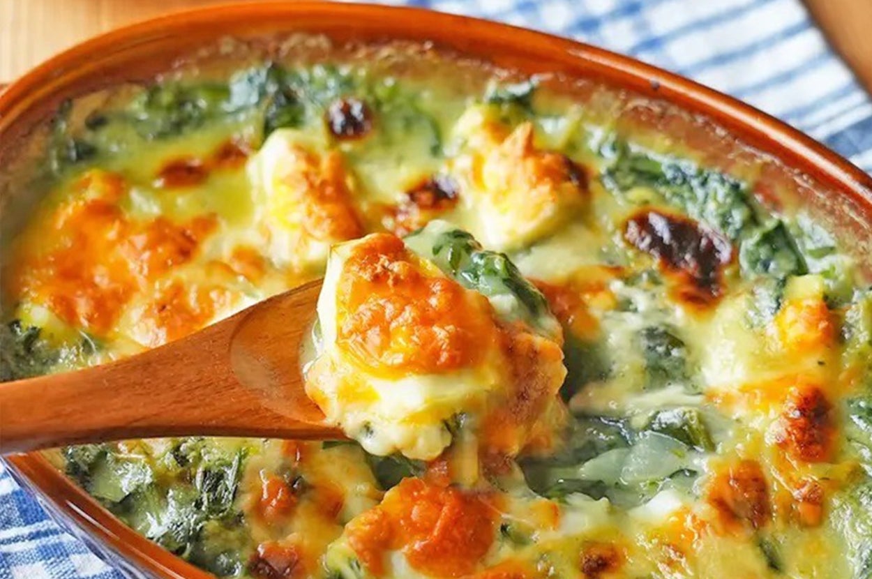 ほうれん草とチーズのグラタンが木のスプーンで盛られている。チェック柄のクロスの上に置かれている。