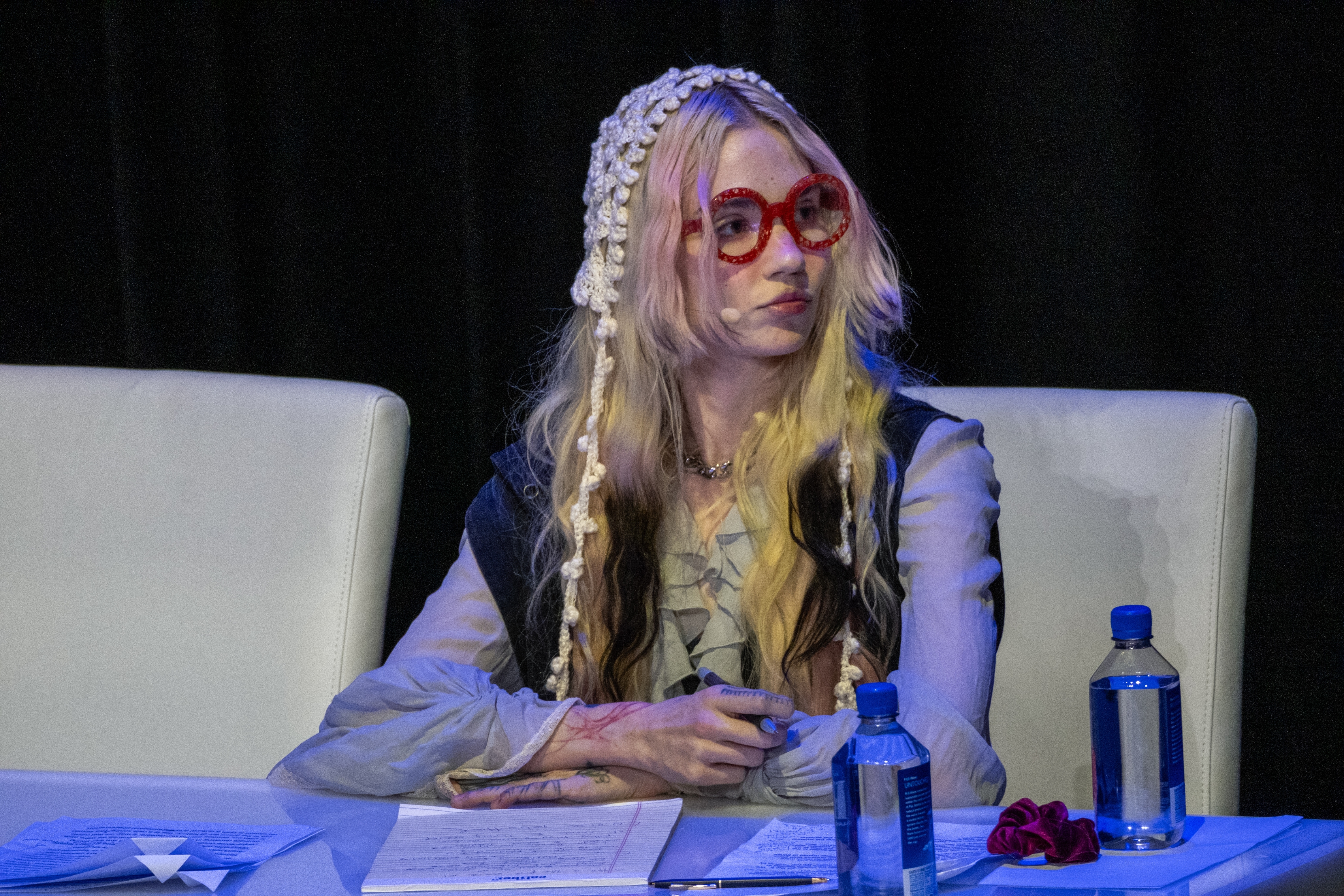 Grimes sits astatine  a array  successful  a ceremonial  setting, wearing reddish  glasses and decorative headwear, with papers and h2o  bottles connected  the table