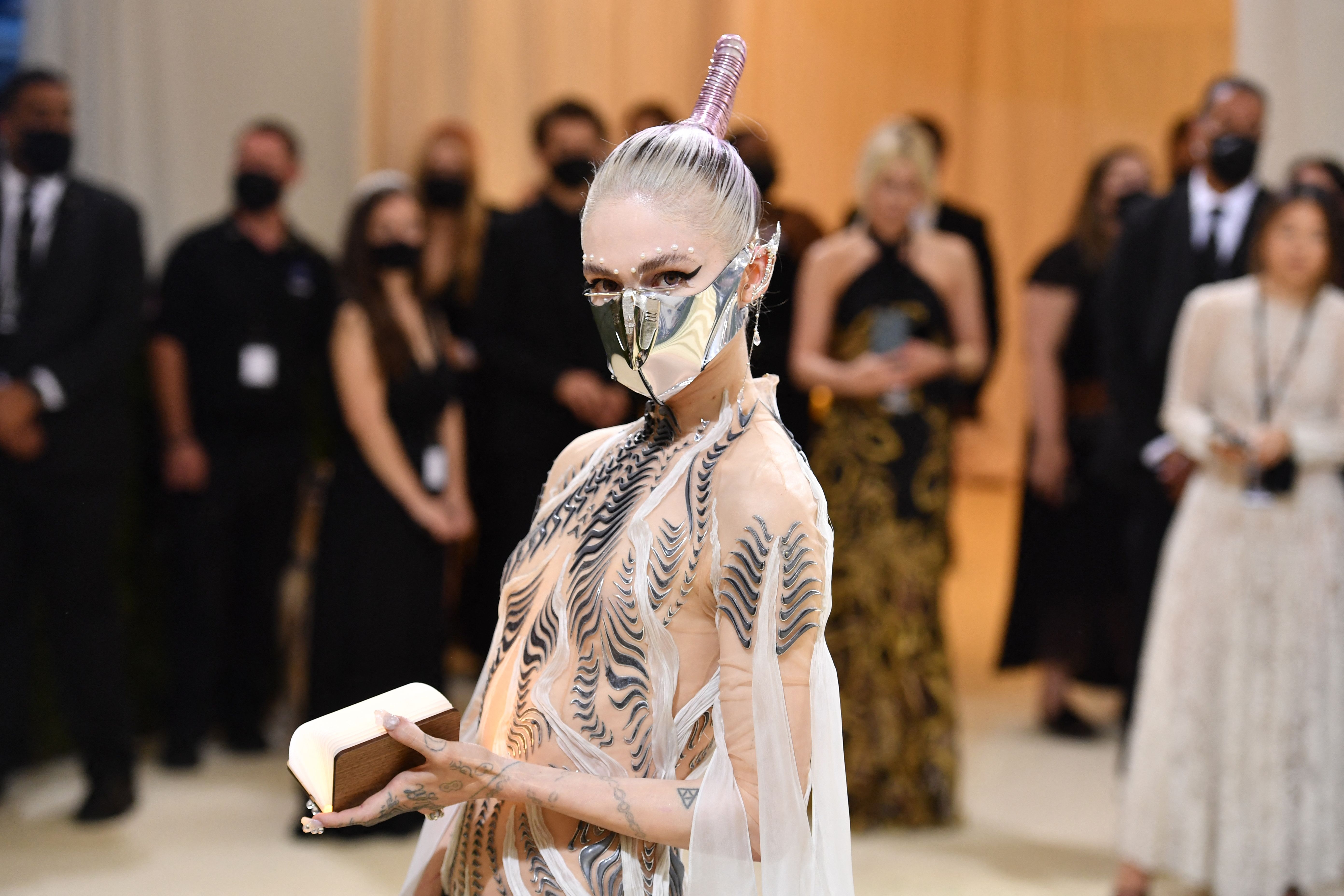 Grimes connected  a reddish  carpet wearing a futuristic gown with look   overgarment   and holding a book, surrounded by masked attendees