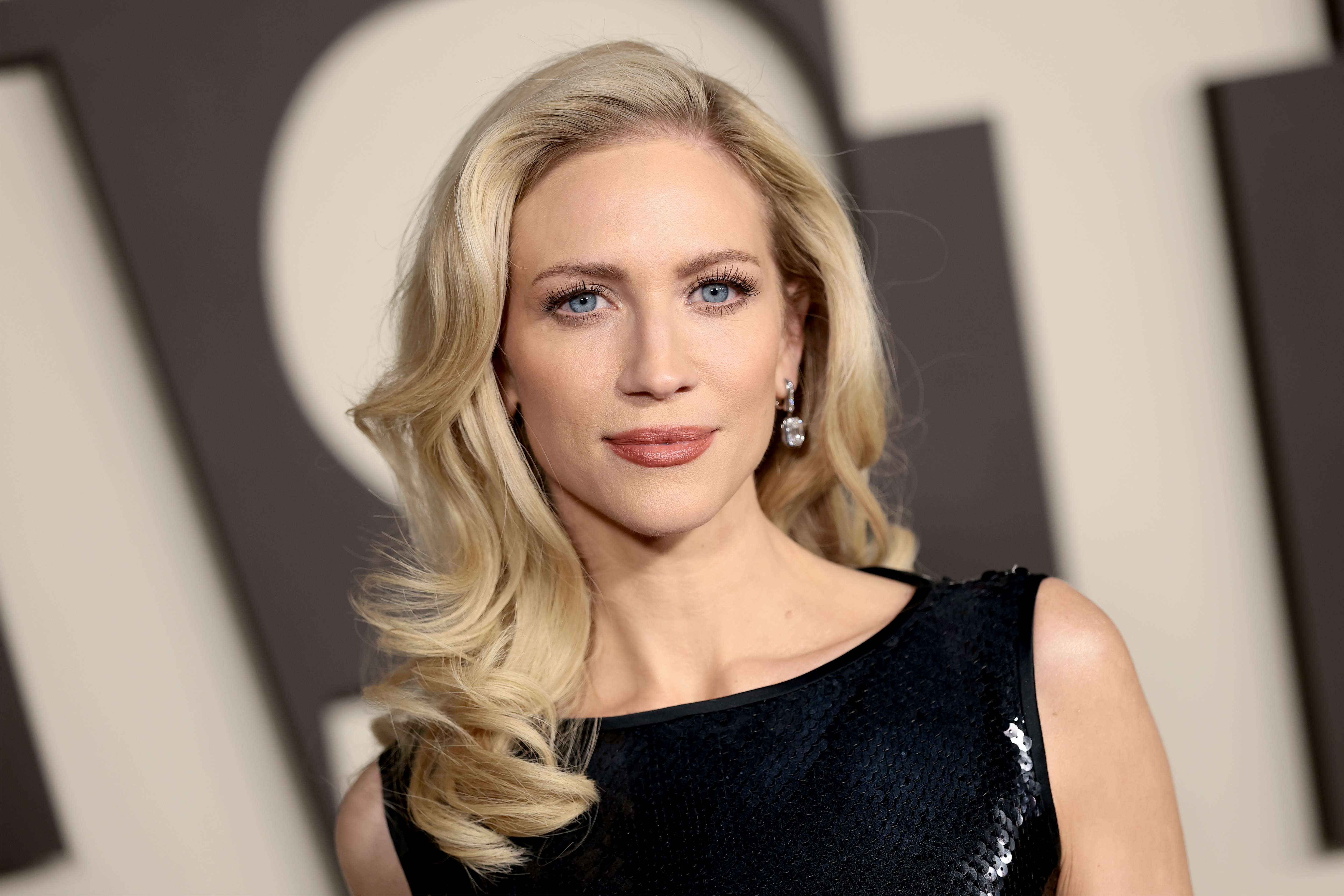 brittany snowfall poses connected a reddish carpet wearing a sleek, sleeveless achromatic formal with sequined details, and elegant earrings