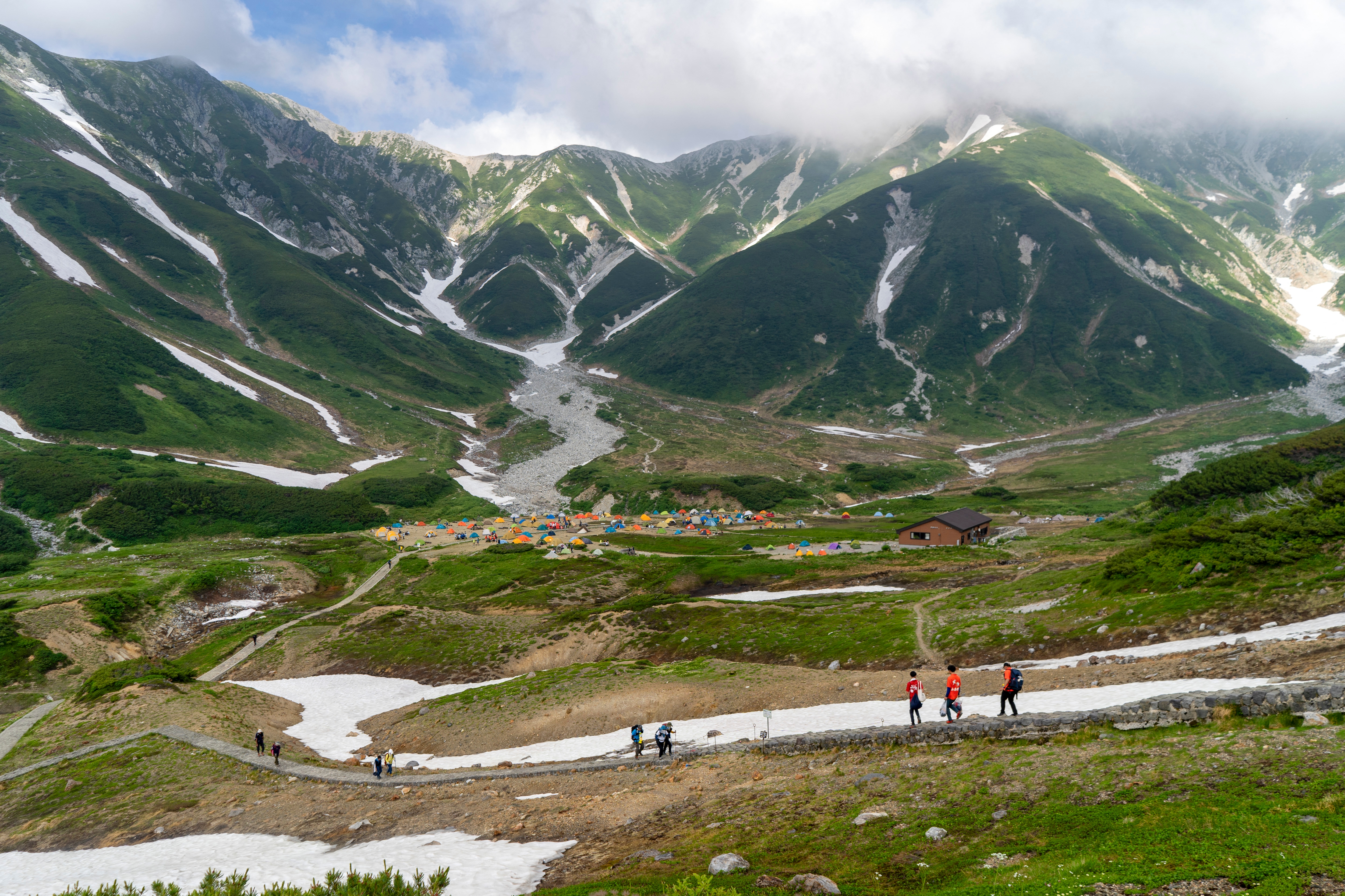Scenic mountain landscape with lush green hills, snow patches, and a campsite with tents in the valley. Hikers walk along a trail in the foreground