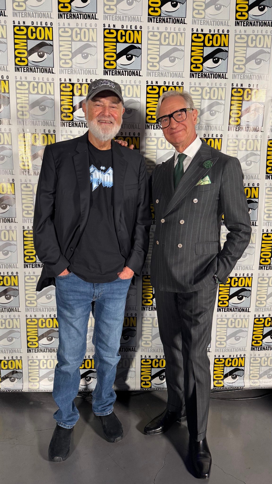 Two men astatine Comic-Con, 1 successful a casual outfit with a denim and t-shirt ensemble, the different successful a tailored pinstripe suit with glasses and a pouch square