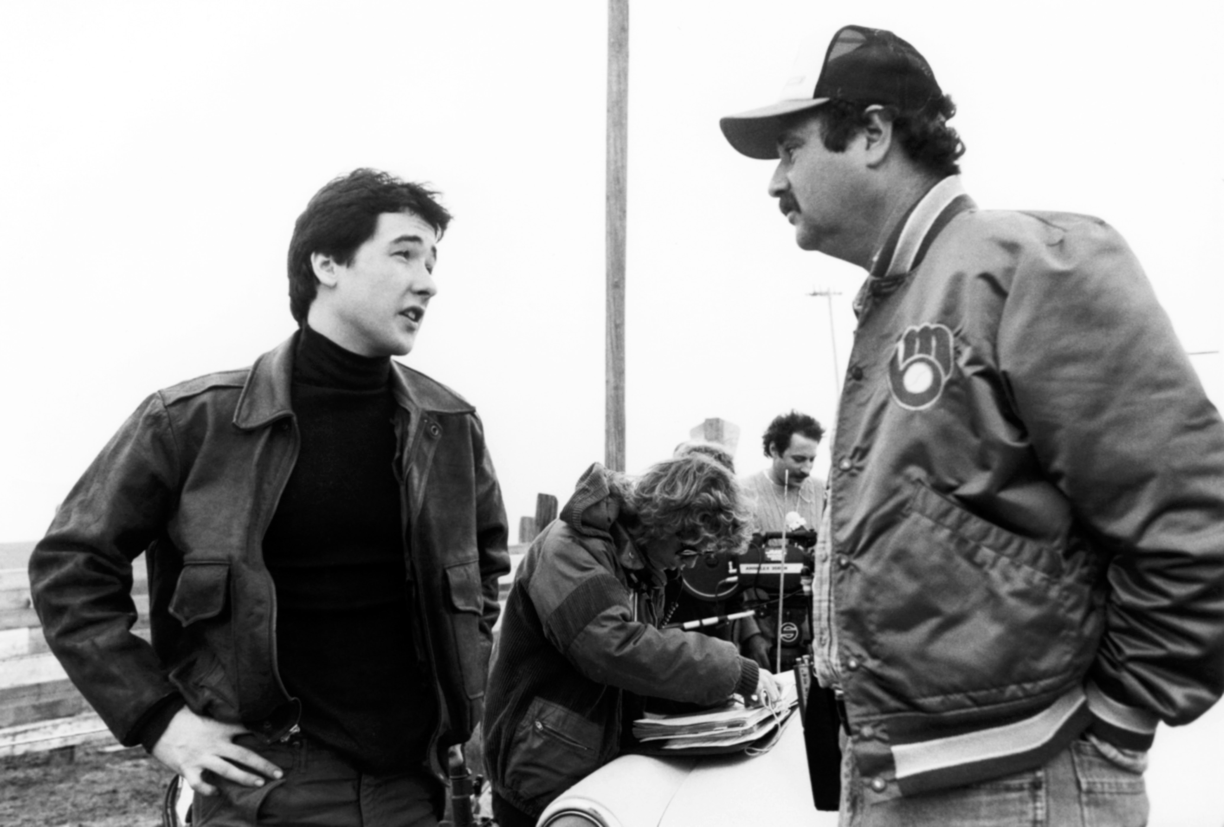 People conversing connected a movie set; 1 wears a casual jacket, the different a headdress and sports jacket. Crew subordinate takes notes successful the background