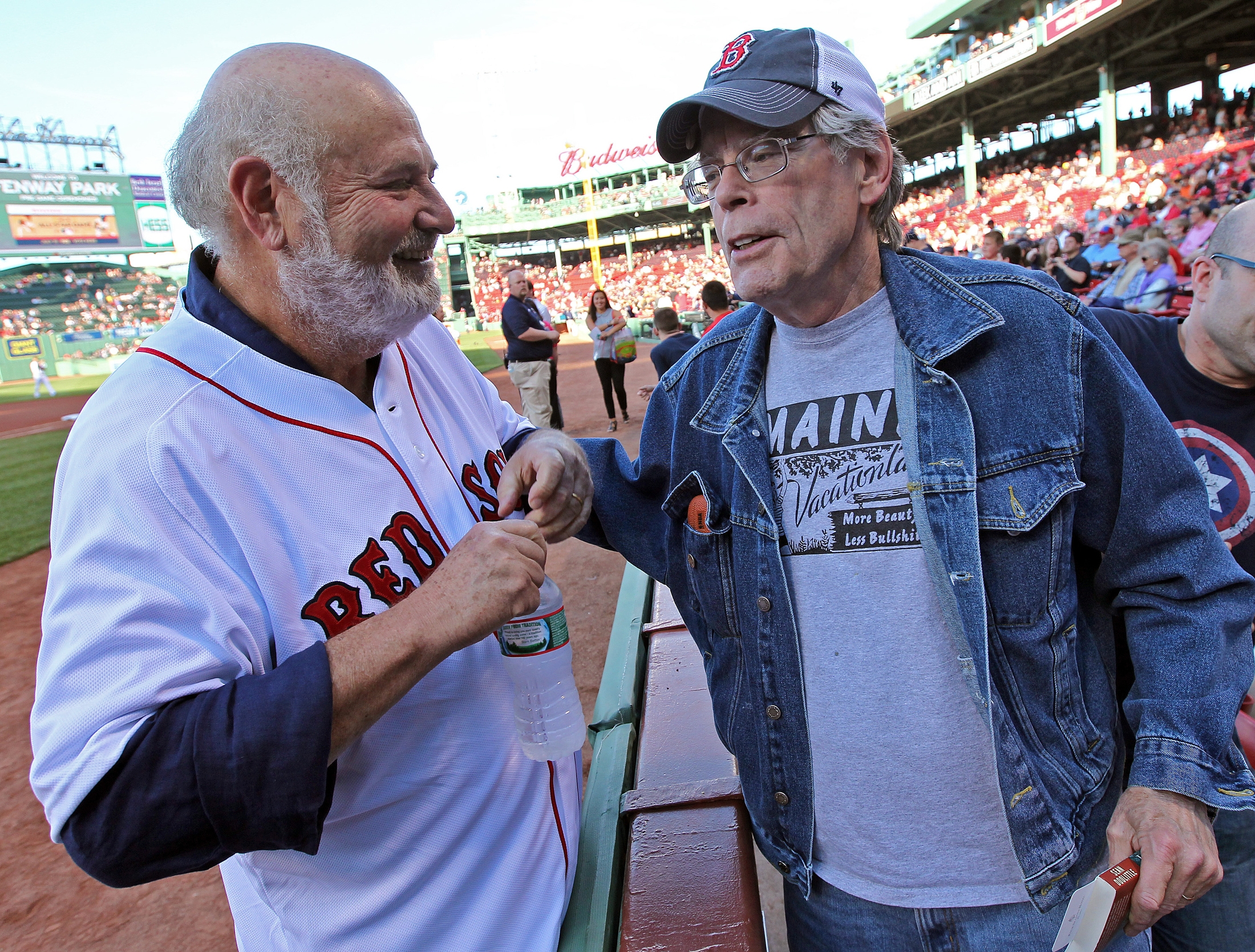 Two older men chat astatine a shot stadium. One wears a sports jersey, portion the different sports a denim overgarment and cap. A crippled is seen successful the background