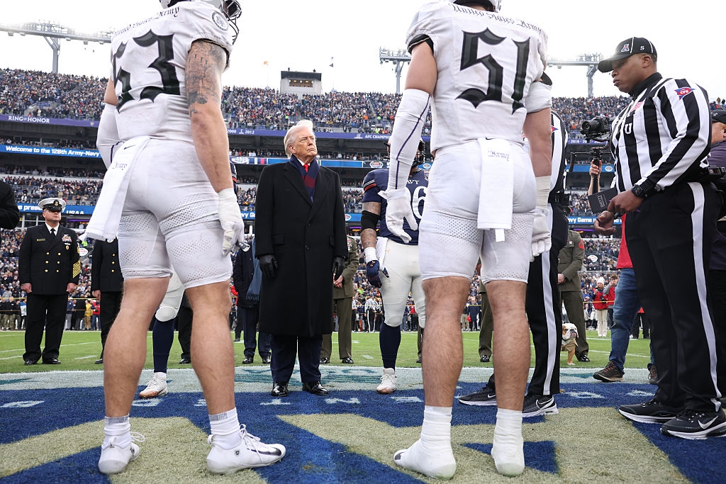 Fotballspillere i uniform samles på midtbanen for en myntkast, med funksjonærer og en bemerkelsesverdig skikkelse iført frakk og skjerf i nærheten. Stadion publikum synlig