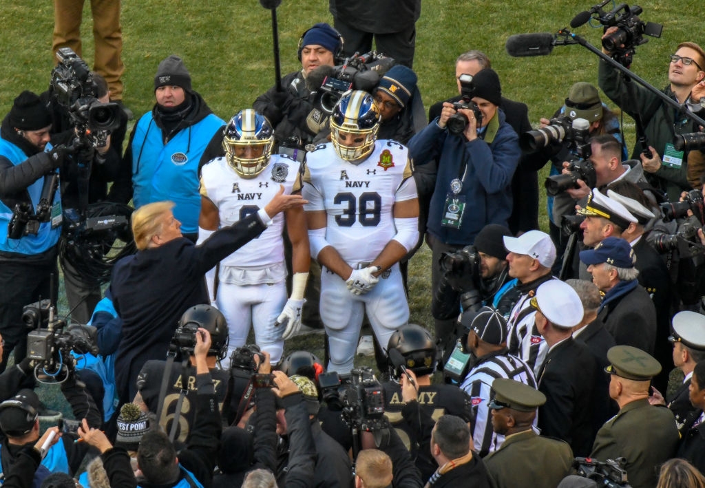En person gestikulerer mot to fotballspillere i marineuniformer, omgitt av fotografer og media, og antyder en pressebegivenhet eller kampatmosfære