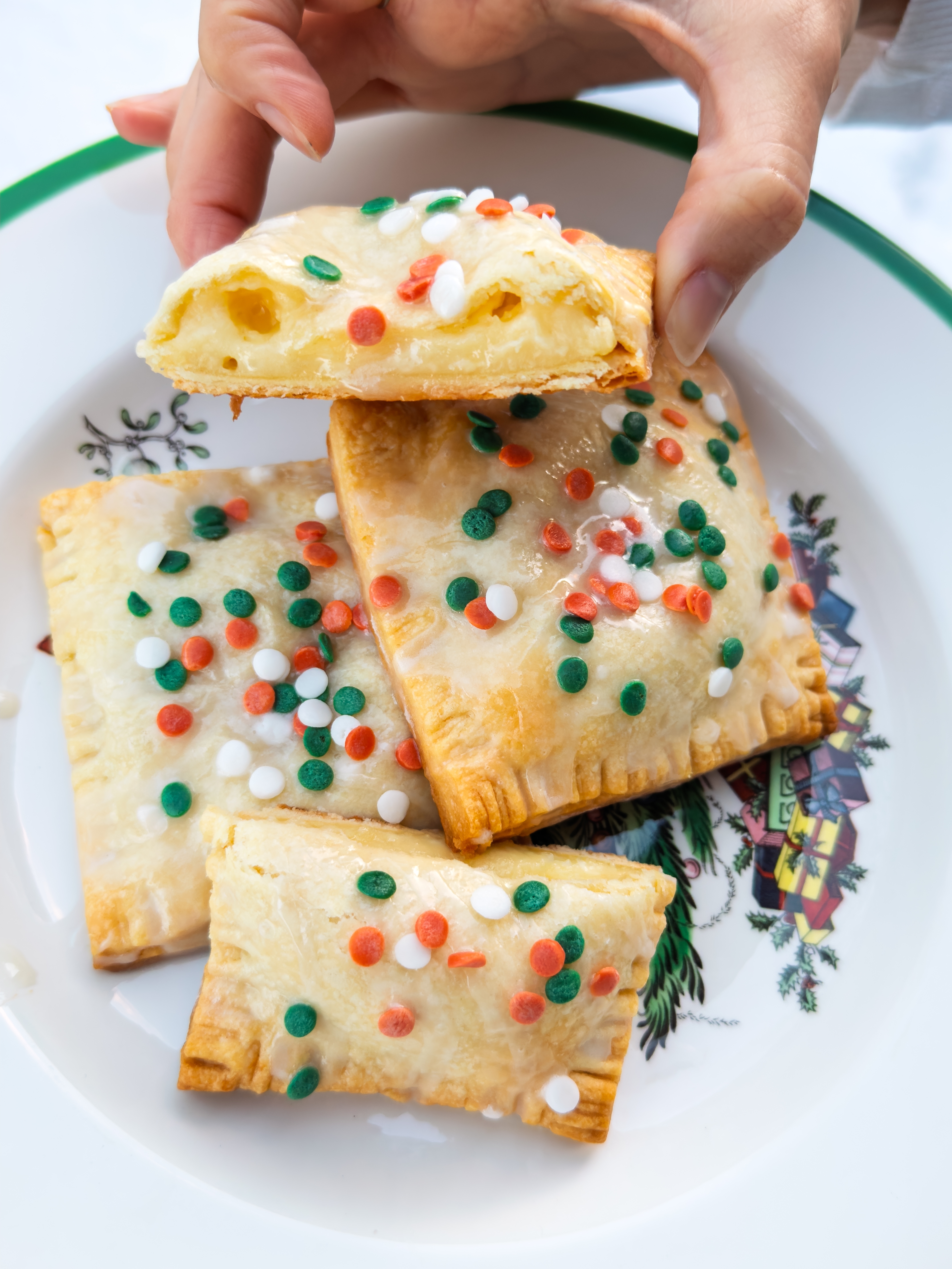 Person holding a flaky pastry with frosting and colorful sprinkles, over a plate with two more pastries