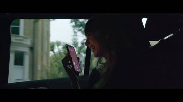 A person smiles while holding a phone to their ear, sitting in a car. The outside view is slightly blurred