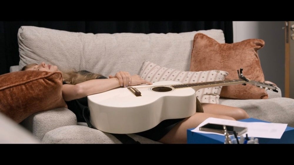 Person resting on a couch holding a guitar, with papers and a phone on a nearby table