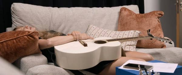 Person resting on a couch holding a guitar, with papers and a phone on a nearby table