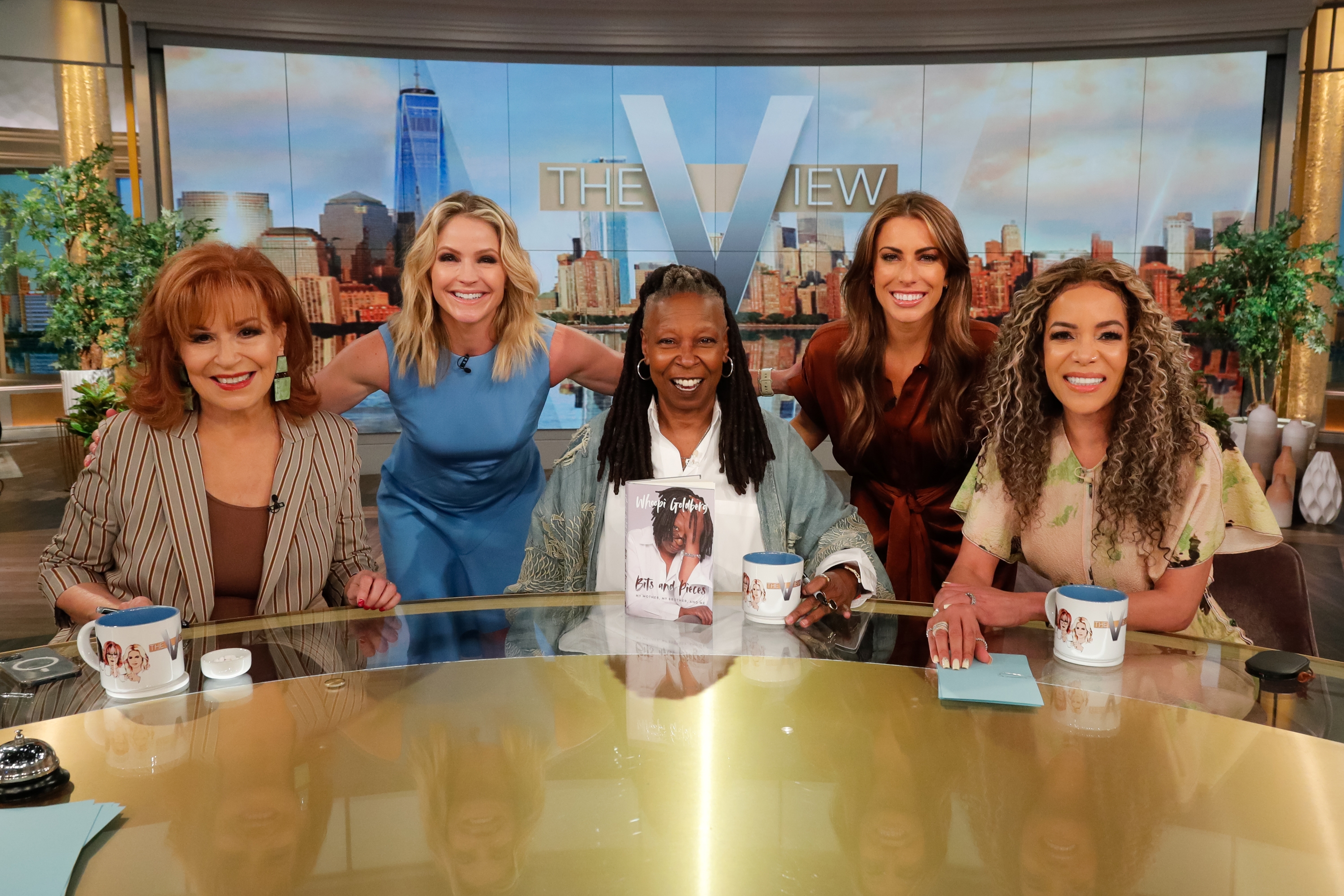 Five women from a speech   amusement   sheet  airs  happily astatine  a array  with mugs and a photograph  book, acceptable   against a cityscape backdrop