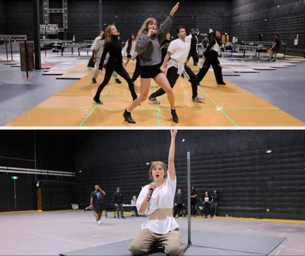 Person leading dance rehearsal in a studio, surrounded by several dancers. They're wearing casual clothing and appear focused on choreography