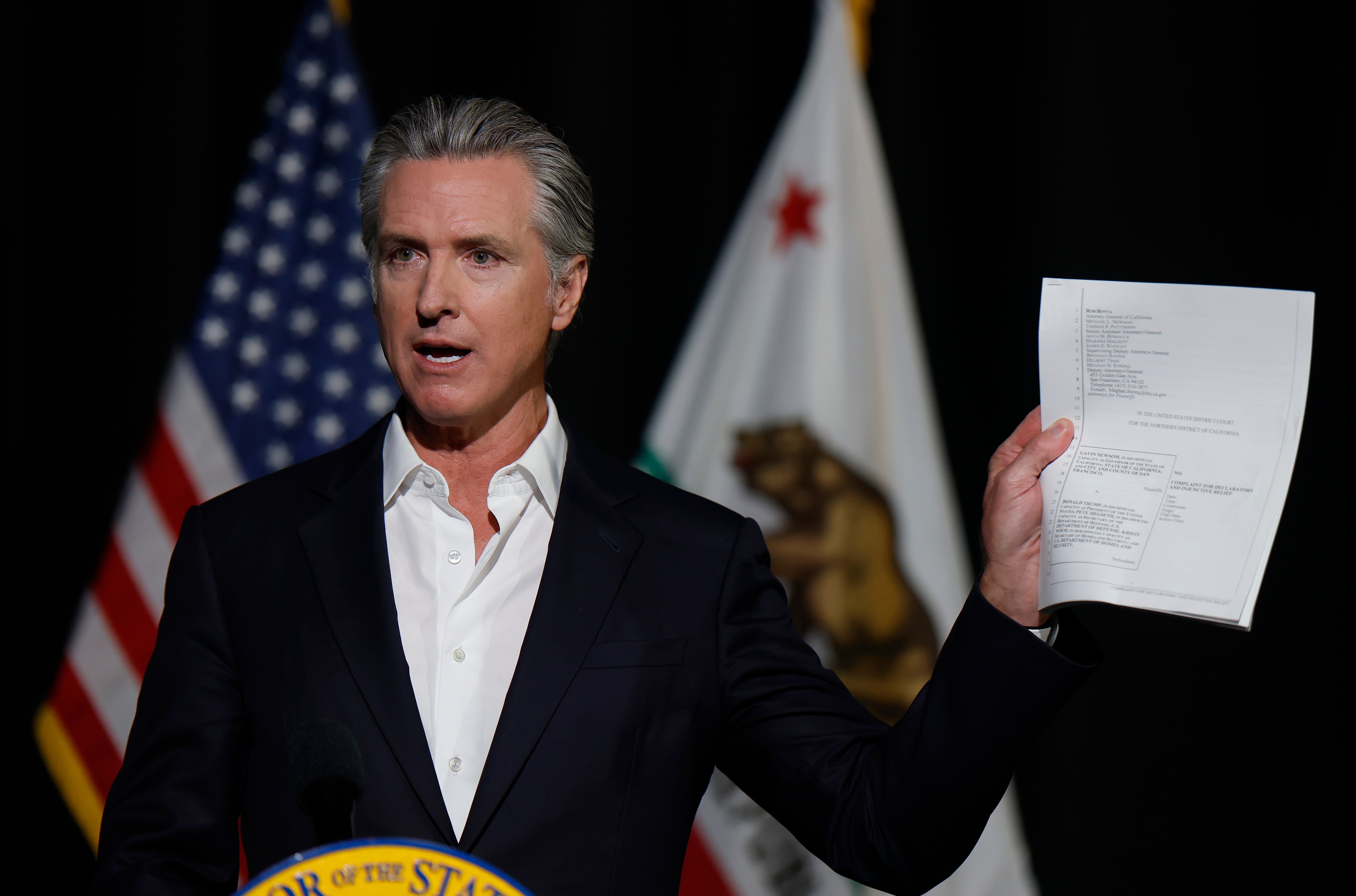 En person i dress snakker ved et podium og holder opp et papir. To amerikanske og California-flagg er synlige i bakgrunnen