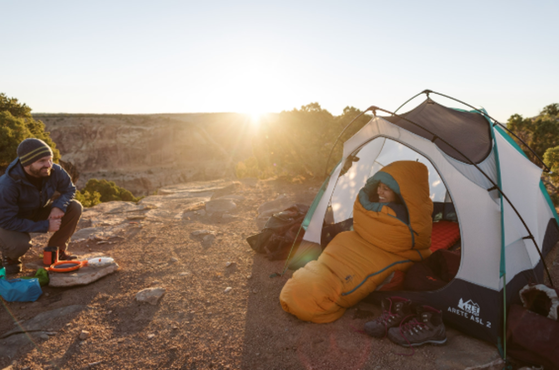 Person sits by a campsite with a structure   astatine  sunrise, different  is wrapped successful  a sleeping bag, illustrating outdoor instrumentality  for camping adventures