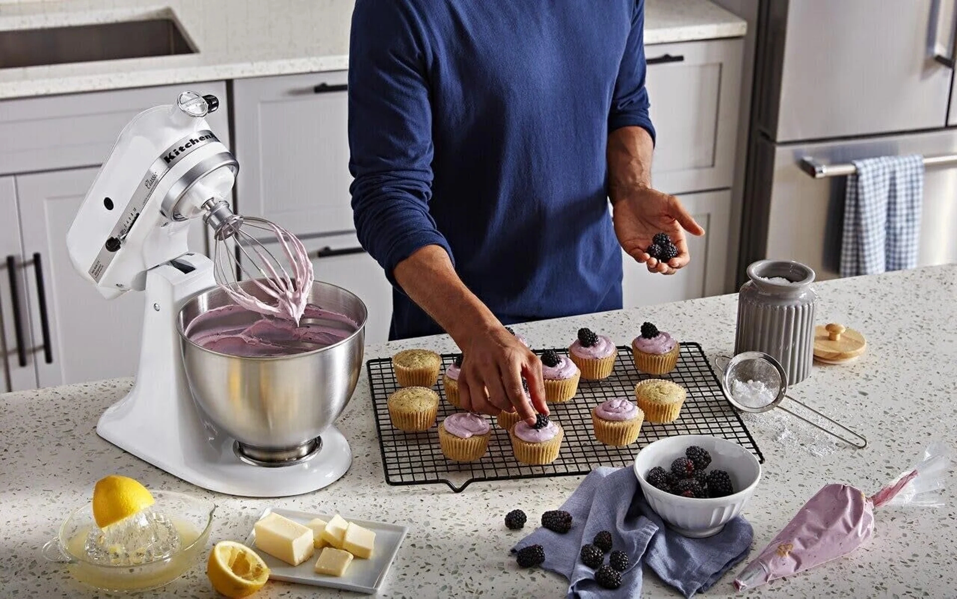 Person icing cupcakes beside a mixer connected  a room  counter; ingredients similar  butter, lemon, and berries are nearby