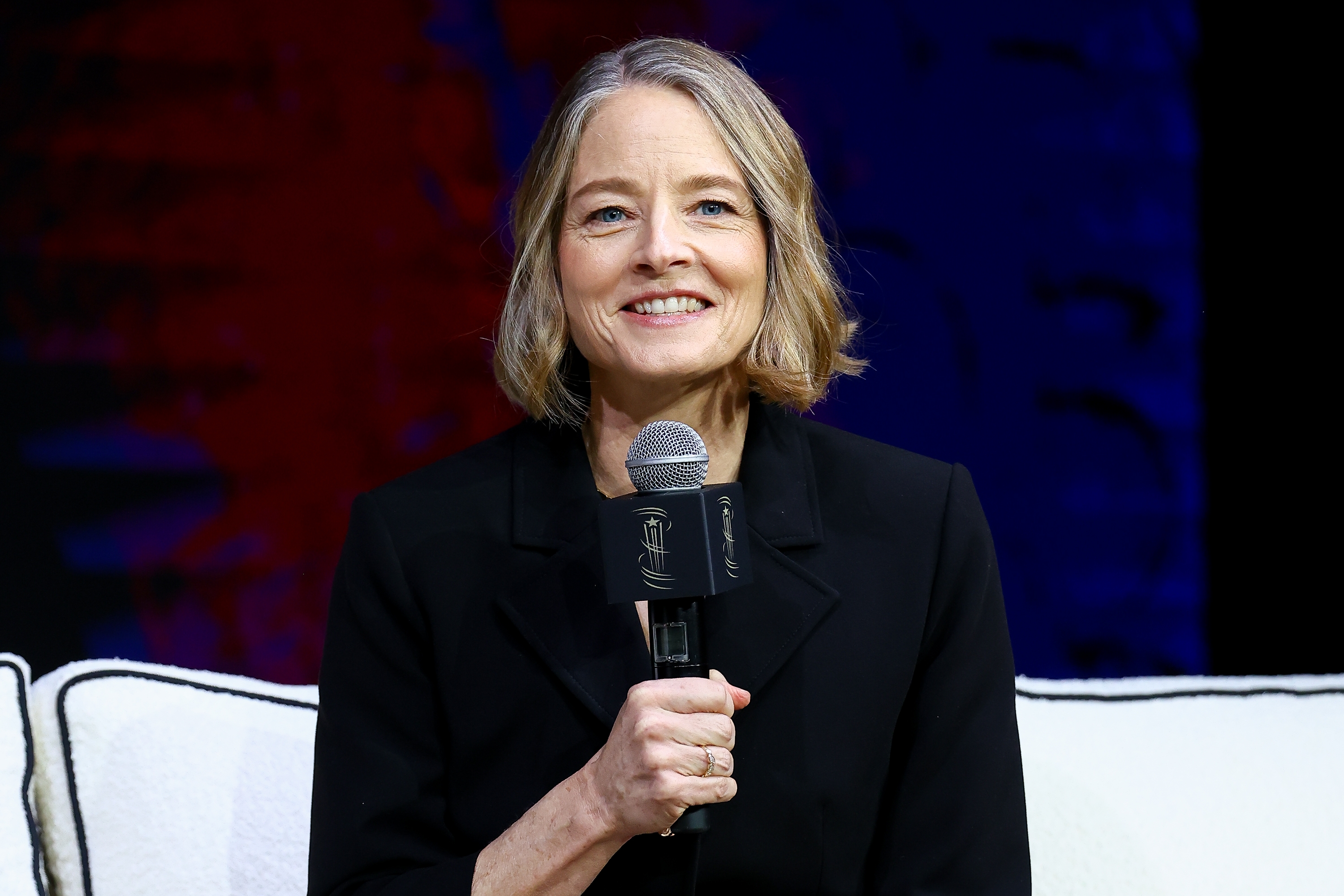 Person holding microphone and smiling, sitting connected  signifier    with a cushioned backdrop, wearing a tailored achromatic  outfit