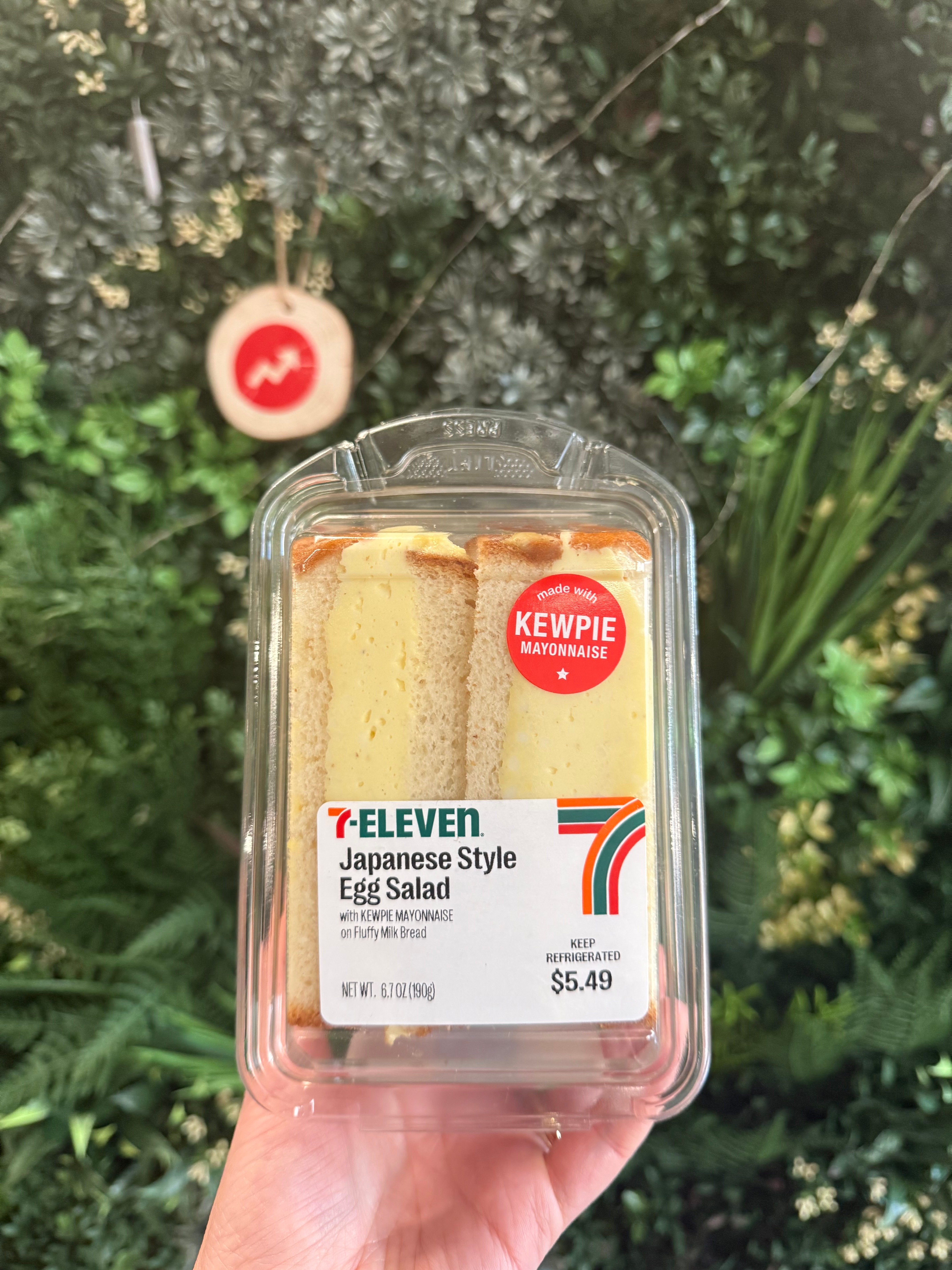 Packaged 7-Eleven Japanese-style egg salad sandwich with Kewpie mayonnaise, held by hand in front of indoor greenery. Price: $5.49