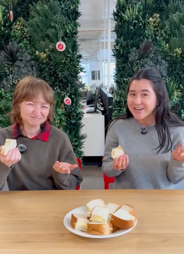 Two people sit at a table, smiling playfully while holding bread. A plate with more bread is in front of them. Lush greenery is in the background