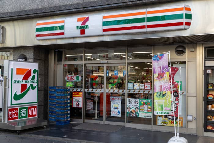 A 7-Eleven storefront with sliding glass doors, displaying promotions and cultural items inside. Signs in Japanese are visible