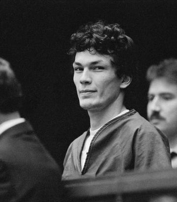 A person in a courtroom looks towards the camera with a slight smile, flanked by a lawyer and an officer in the background