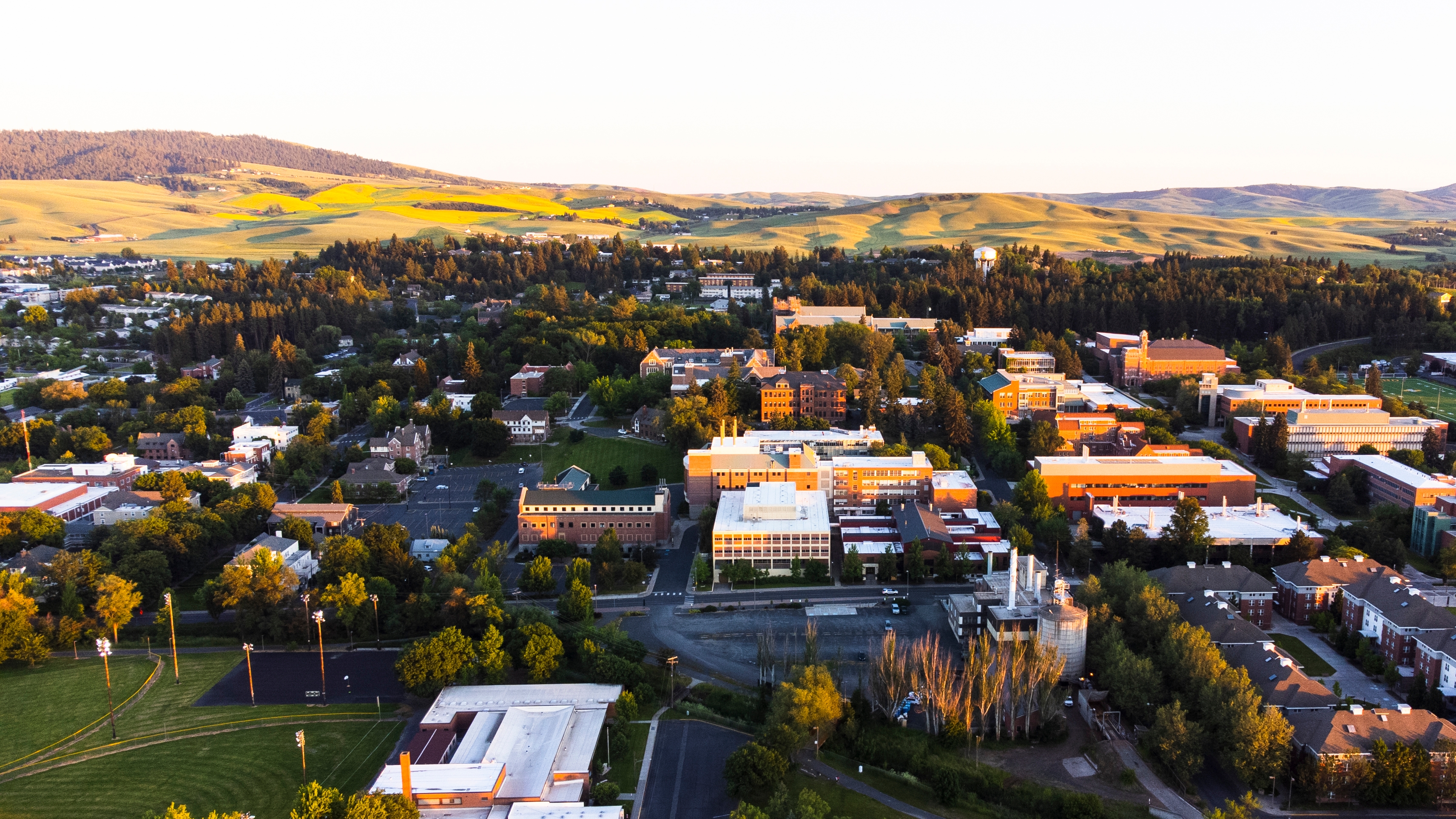 Luftfoto av et universitetscampus omgitt av åser og jorder, med mange bygninger og grønne områder, under en klar himmel